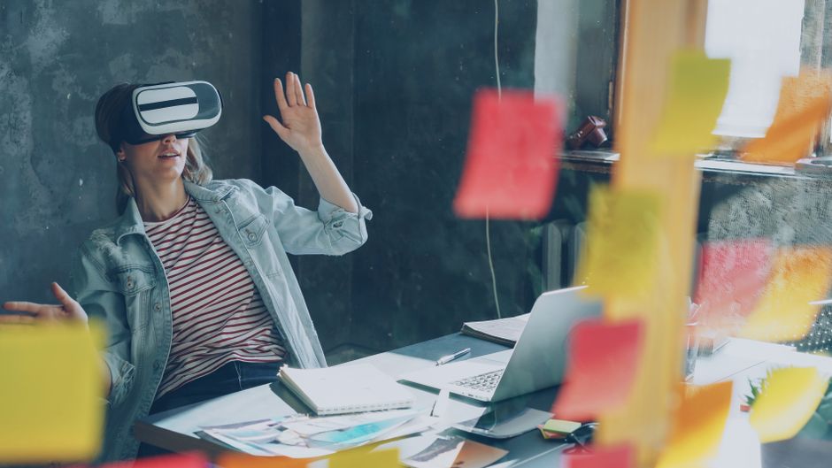 Žena koristi VR headset za radnim stolom uz bilješke i laptop